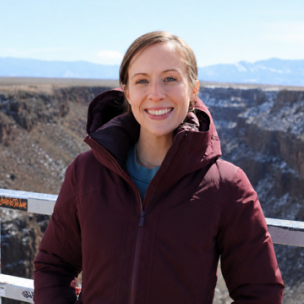 Smiling woman in a burgundy winter jacket at an outdoor overlook, with a canyon and snow-capped mountains in the background.