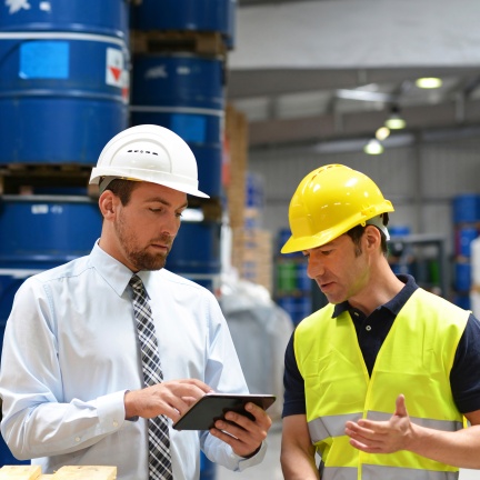 Two men in a warehouse discuss data on a tablet: one wearing a white hard hat and tie, the other in a yellow safety vest and helmet.
